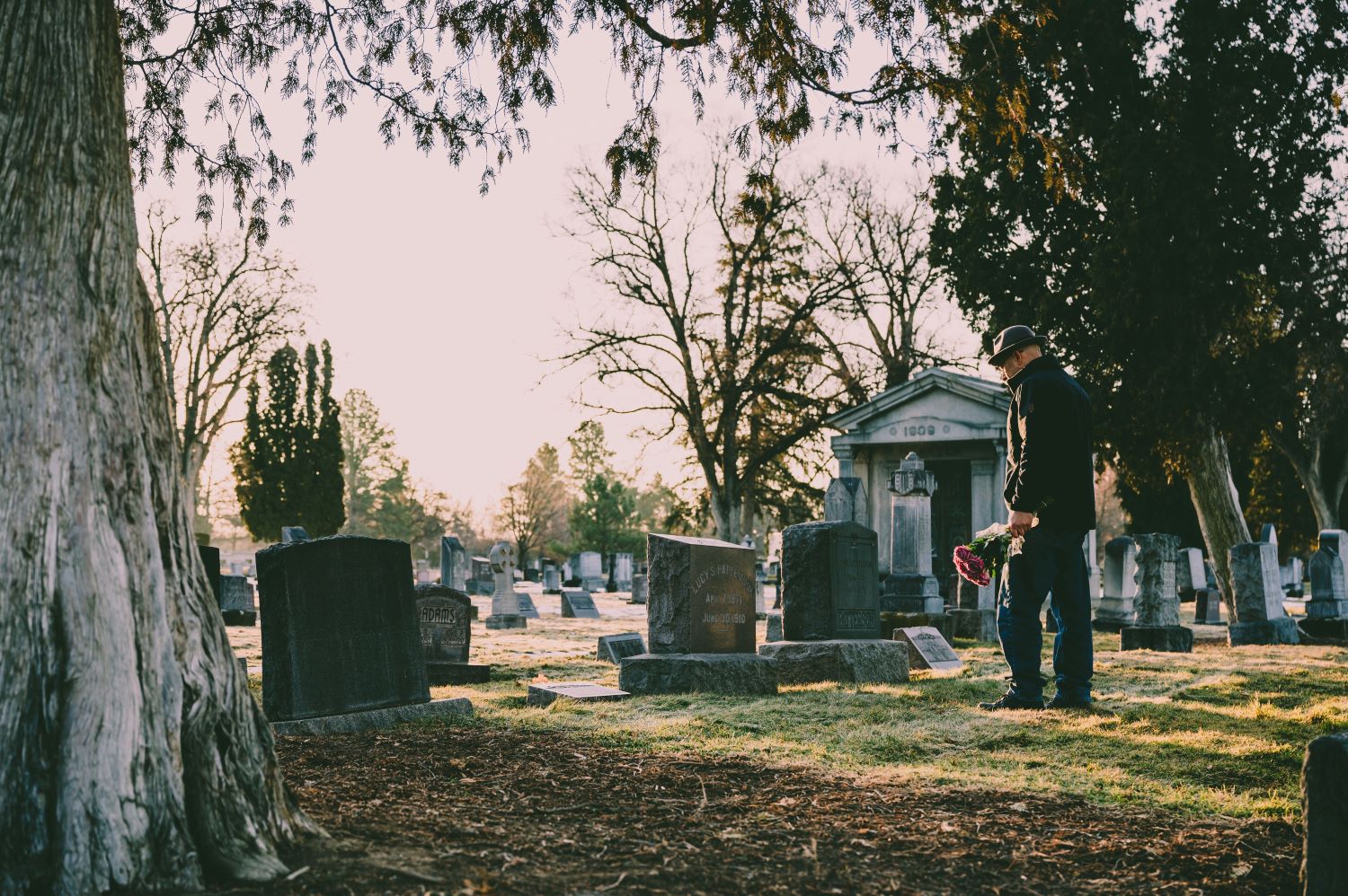 image of a man standing over a headstone in a cemetery with flowers in his left hand